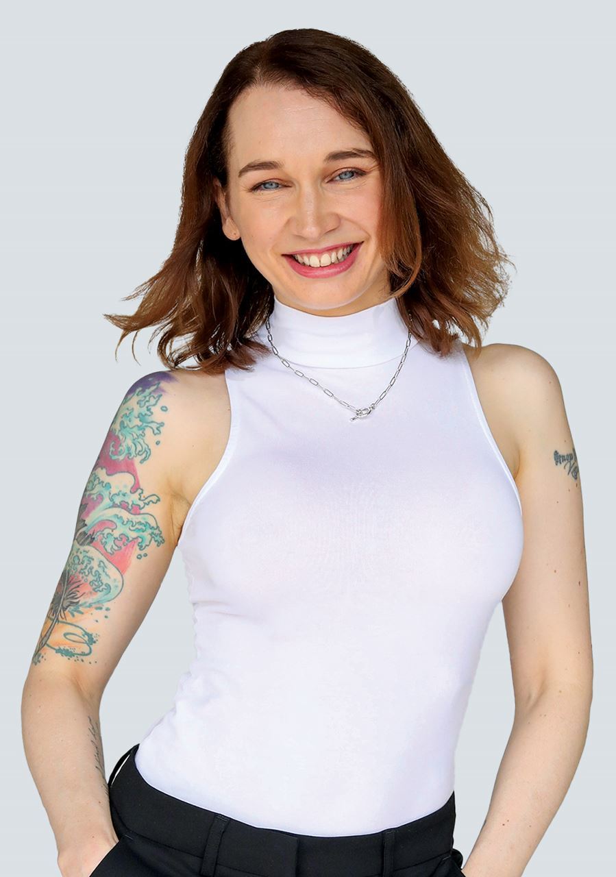 An image of Flic on the left of a white tile. She has auburn hair, a white turtleneck top, silver necklace and a tattoo on her right arm. She is smiling at the camera.